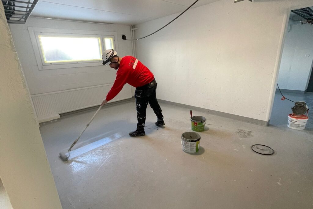 Maalari levittää telalla harmaata maalia betonilattialle punainen paita yllään