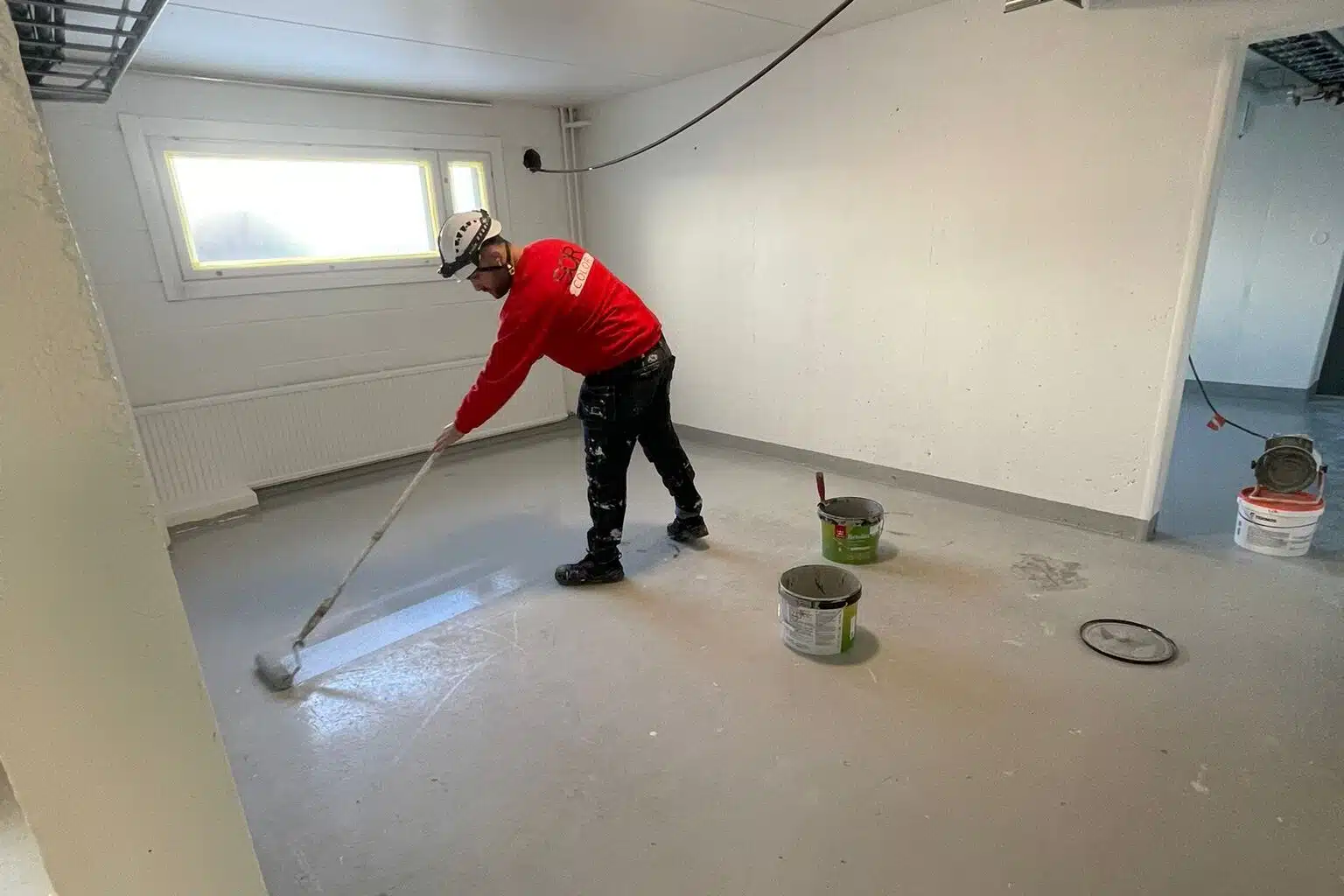 Maalari levittää telalla harmaata maalia betonilattialle punainen paita yllään