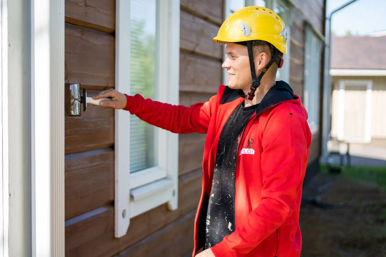 Mies maalaa hirsiseinää punainen paita yllään