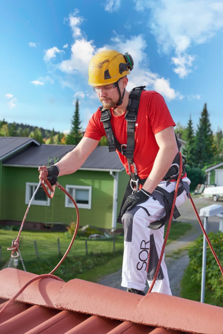 Katon maalaaja levittää maalia pirkävartisella ruiskulla tiilikaton pintaan työvaattteet yllään
