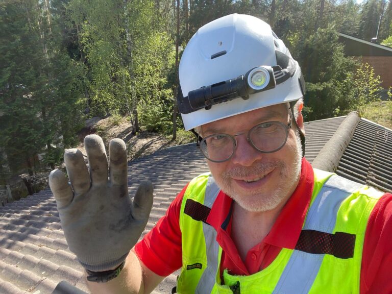 Hymyilevä työmies nostaa kättä kameralle taustalla tiilikatto