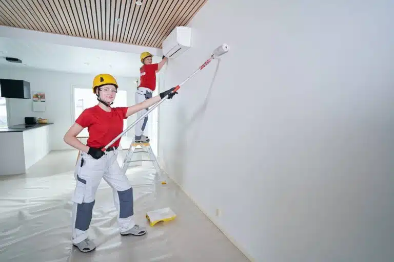 Kaksi maalaria levittävät valkoista maalia sisäseinään hymyillen