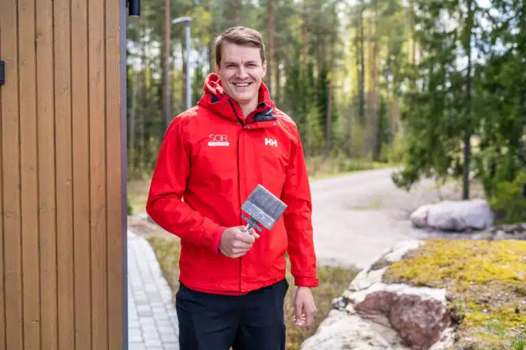 Mies seisoo talon seinän vieressä pensseli kädessä, punainen takki yllään. kuvastaa syksyllä maalaamista