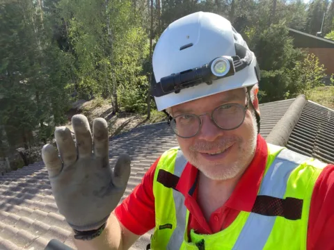 Hymyilevä työmies nostaa kättä kameralle taustalla tiilikatto|||