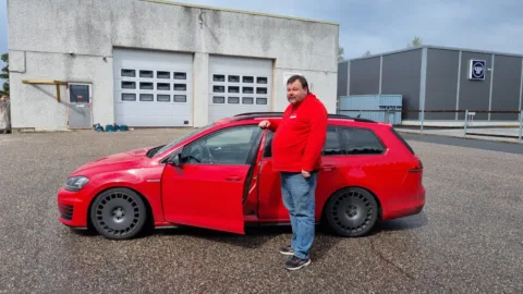 Mien punaisessa paidassa seisoo punaisen auton edessä ja hymyilee||