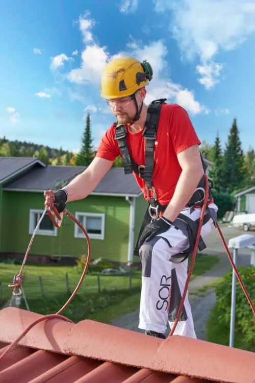 Katon maalaaja levittää maalia pirkävartisella ruiskulla tiilikaton pintaan työvaattteet yllään|