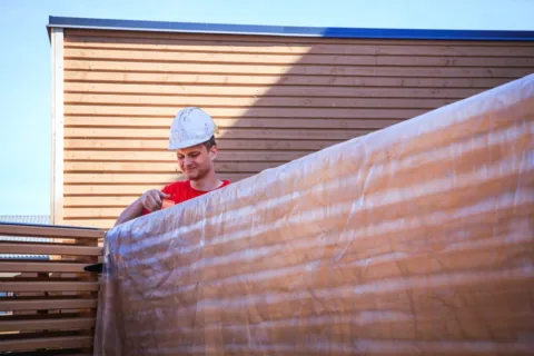 maalari maalaa aitaa punainen paita päällään|||Keltainen talo juuri maalattuna metsän keskellä auringon valaisemana|
